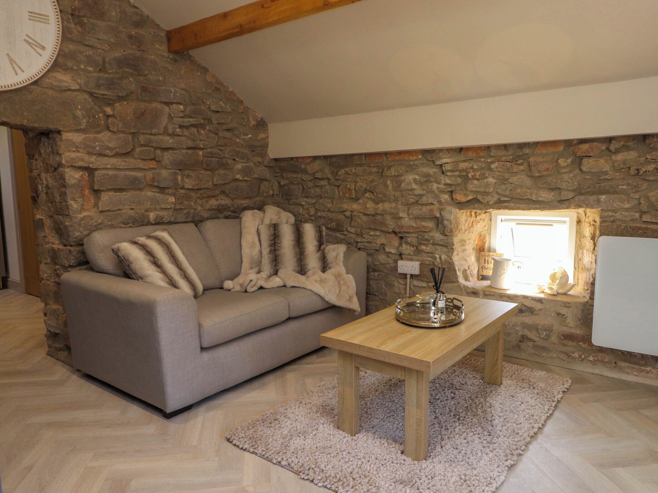 A cozy room with a stone wall, a grey sofa with fur throws, a wooden coffee table, and a small window with a view.