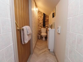 Un pasillo estrecho con una puerta de madera, una toalla blanca colgada del pomo, una pared de piedra, un inodoro blanco y un lavabo blanco con un espejo encima.