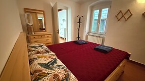 Ein Schlafzimmer mit einem hölzernen Bettgestell, einer roten Bettdecke und einem floralen Kissen. Es gibt einen Schminktisch mit Spiegel, einen Kleiderständer und ein Fenster mit Fensterläden.