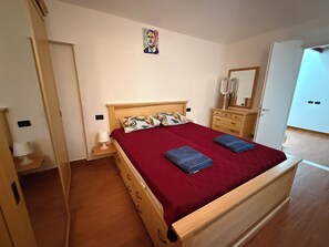 A bedroom with a wooden bed, a red bedspread, two pillows, a nightstand, a mirror, and a door leading to another room.