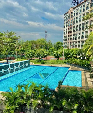 A swimming pool with clear blue water, surrounded by lush greenery and a tall building in the background. - IV’s Condo with AC, WiFi, Netflix in Cainta (Cainta)