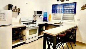 A compact kitchen with a stove, oven, microwave, and sink. There are three stools at the counter, and a blue curtain hangs over the window.