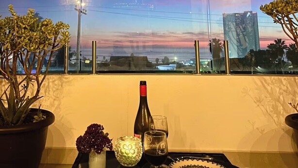 A balcony with a table set for two, featuring a bottle and glasses, potted plants, and a decorative glass.