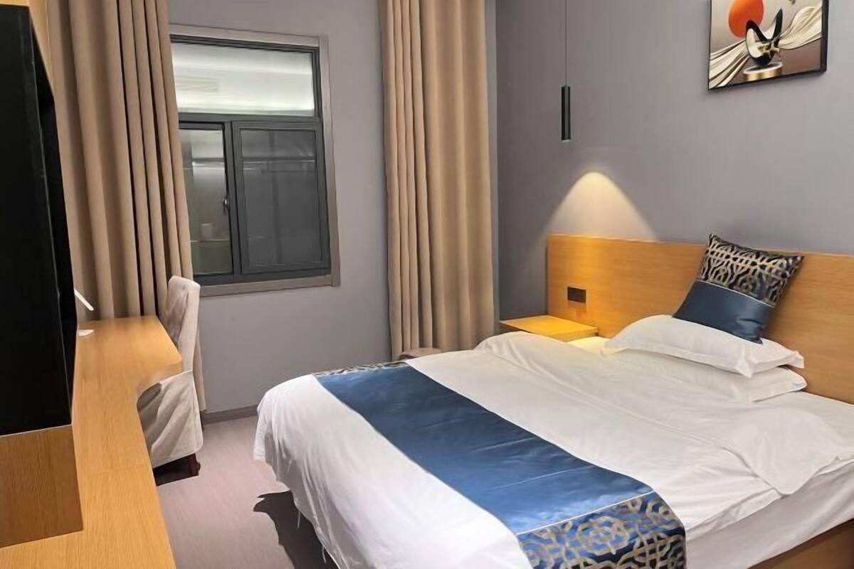 A hotel room with a bed, a nightstand, a lamp, a window with curtains, and a painting on the wall.