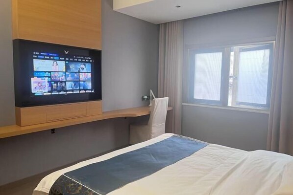A hotel room with a bed, a TV mounted on the wall, a window with curtains, and a wooden shelf.