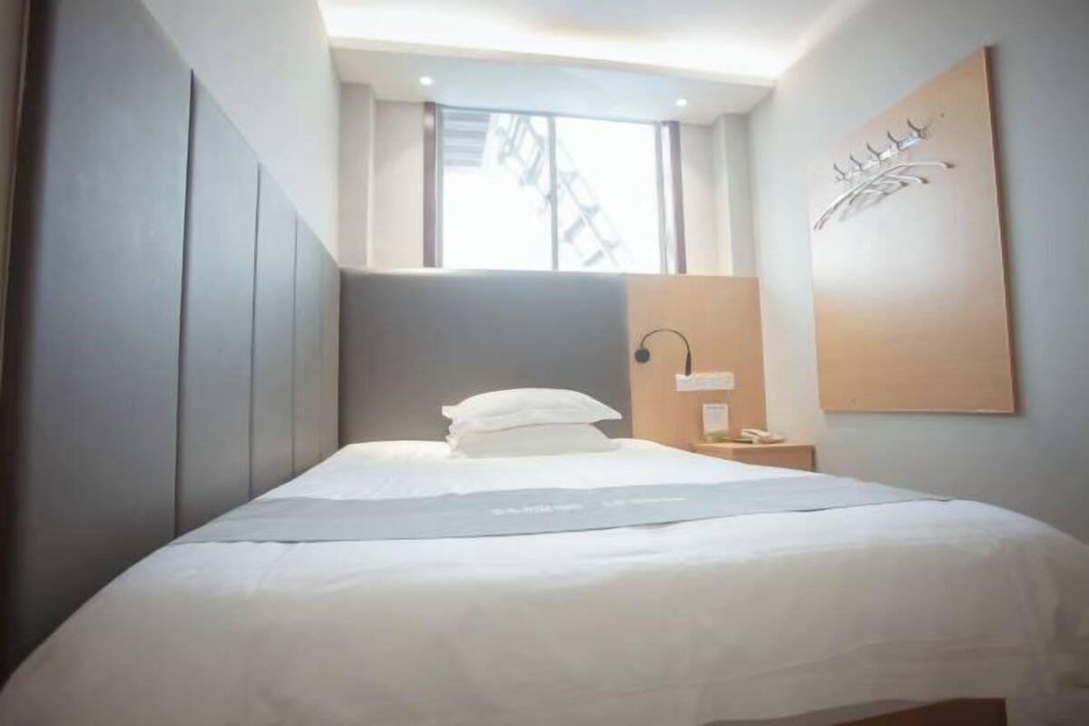 A modern hotel room with a bed, a nightstand, a wall-mounted lamp, and a window with a view of a staircase.