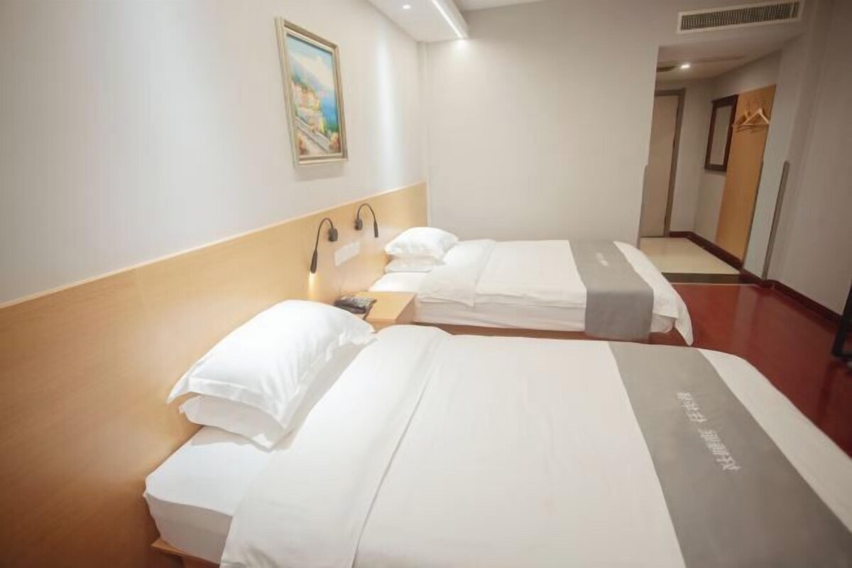 Une chambre d’hôtel avec deux lits, chacun agrémenté de linge de lit blanc et d’un élément décoratif gris. Un tableau est accroché au mur au-dessus des lits, et un porte-manteau se trouve dans un coin de la pièce.