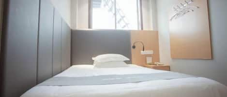 A modern hotel room with a bed, a nightstand, a wall-mounted lamp, and a window with a view of a building outside.
