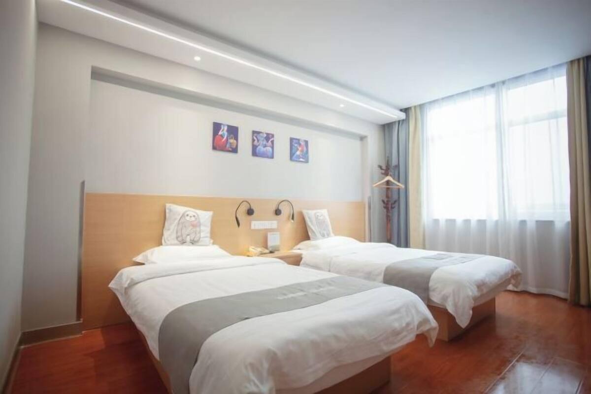 A hotel room with two beds, a wooden headboard, a bedside table with a lamp, and a window with curtains.