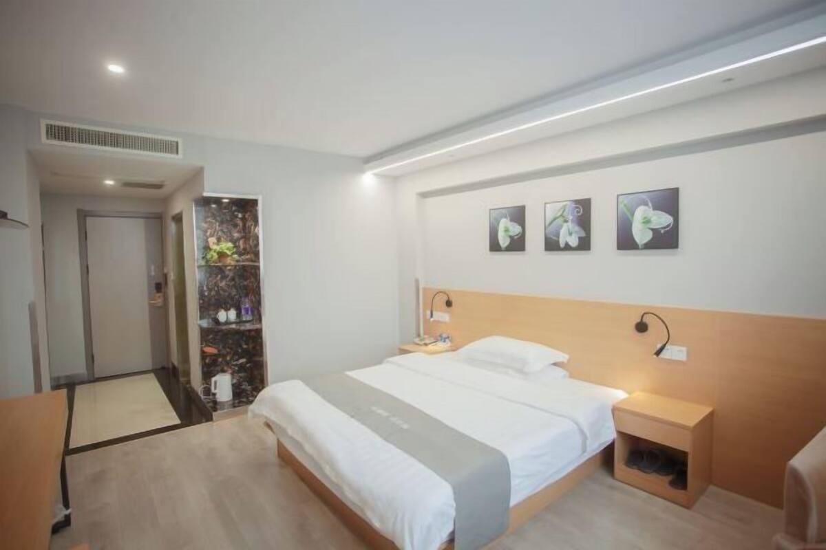 A modern hotel room with a bed, bedside tables, a nightstand, a wall-mounted lamp, and framed artwork.