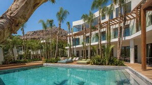 Zona de piscina con una piscina azul cristalina, rodeada de palmeras y un edificio moderno con varios balcones.