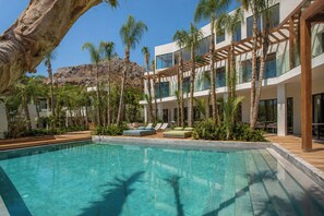 Zona de piscina con una piscina azul cristalina, rodeada de palmeras y un edificio moderno con varios balcones.