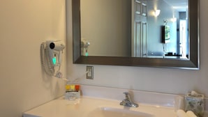A bathroom with a large mirror, a white sink, a hairdryer, and a towel dispenser. - Kow's Inn (Bras D'or)