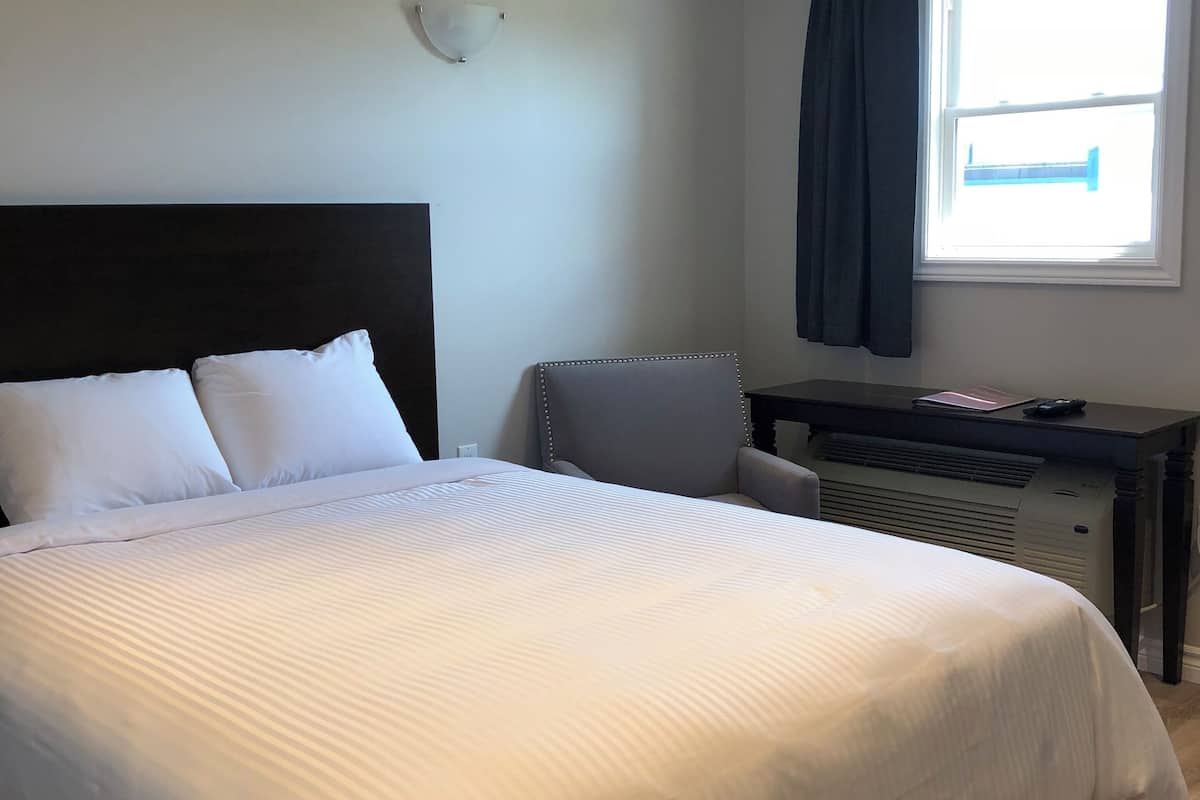 A neatly made bed with white linens, a dark headboard, a small desk with a chair, and a window with curtains.