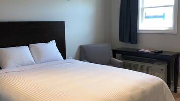 A neatly made bed with white linens, a dark headboard, a small desk with a chair, and a window with curtains.