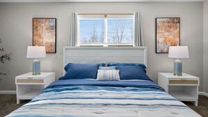 A bedroom with a large bed, two bedside tables with lamps, a window with blinds, and two abstract paintings on the walls.