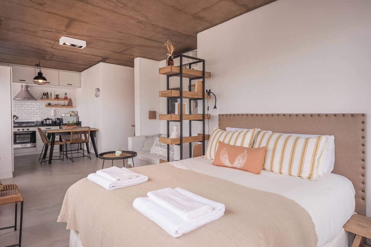 A modern hotel room with a bed, a small dining area, and a kitchenette.