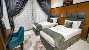 A hotel room with two beds, a desk, a blue chair, and a large window with curtains. - Grand Season 3 (Al Khobar)