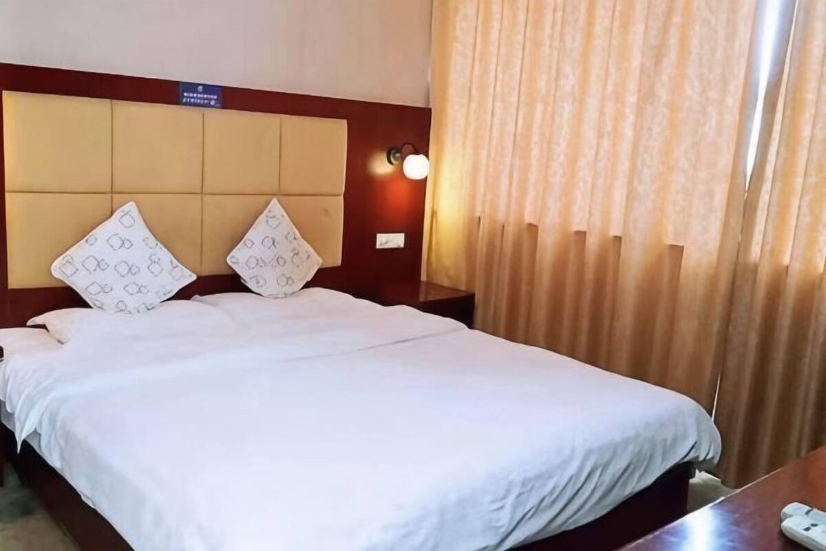 Quarto de hotel com cama, duas almofadas, cabeceira, mesa de cabeceira, abajur e cortina.