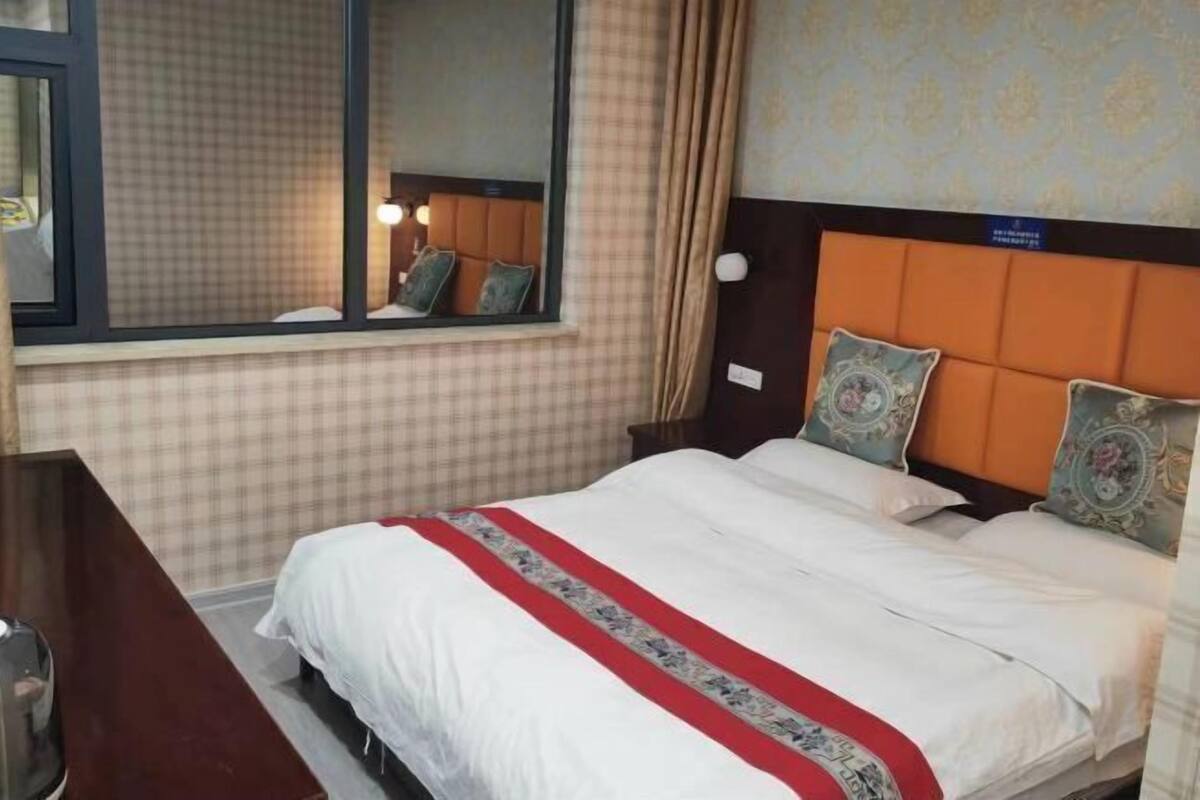 Quarto de hotel com uma cama grande, uma janela com cortinas, cabeceira com almofadas decorativas e parede com padrão xadrez.