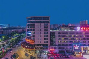 Un edificio de varios pisos con fachada curva, un estacionamiento y edificios comerciales adyacentes con señalización iluminada.