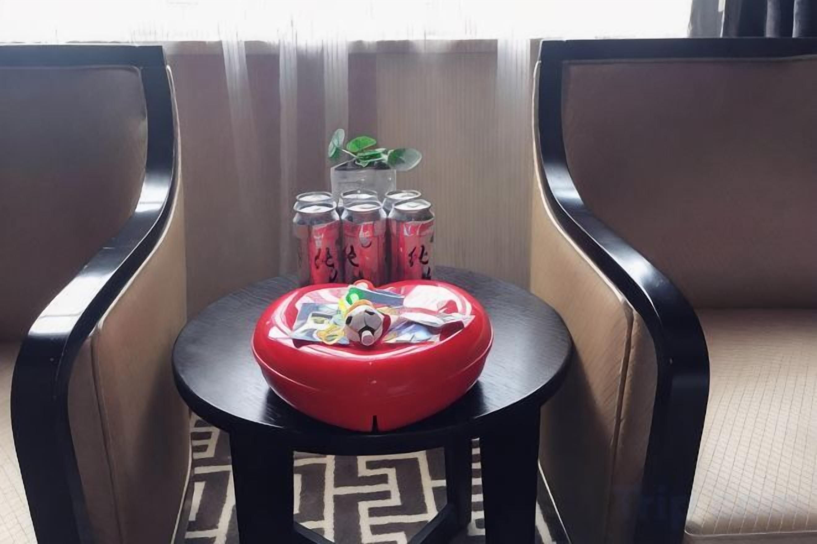 A small round table with a red bowl, a toy, and a plant. Behind the table, there are two chairs and a window with curtains.
