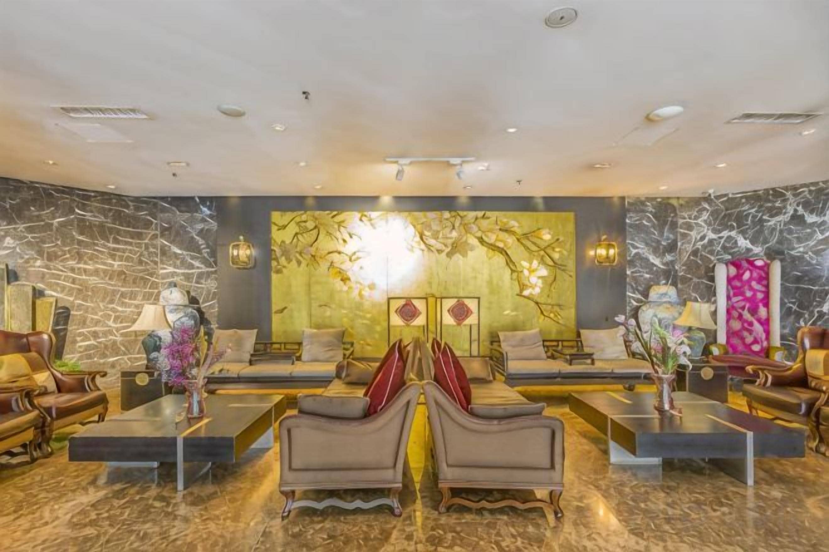 A spacious lobby with a marble floor, a central seating area with sofas and armchairs, and a decorative wall with a golden mural.