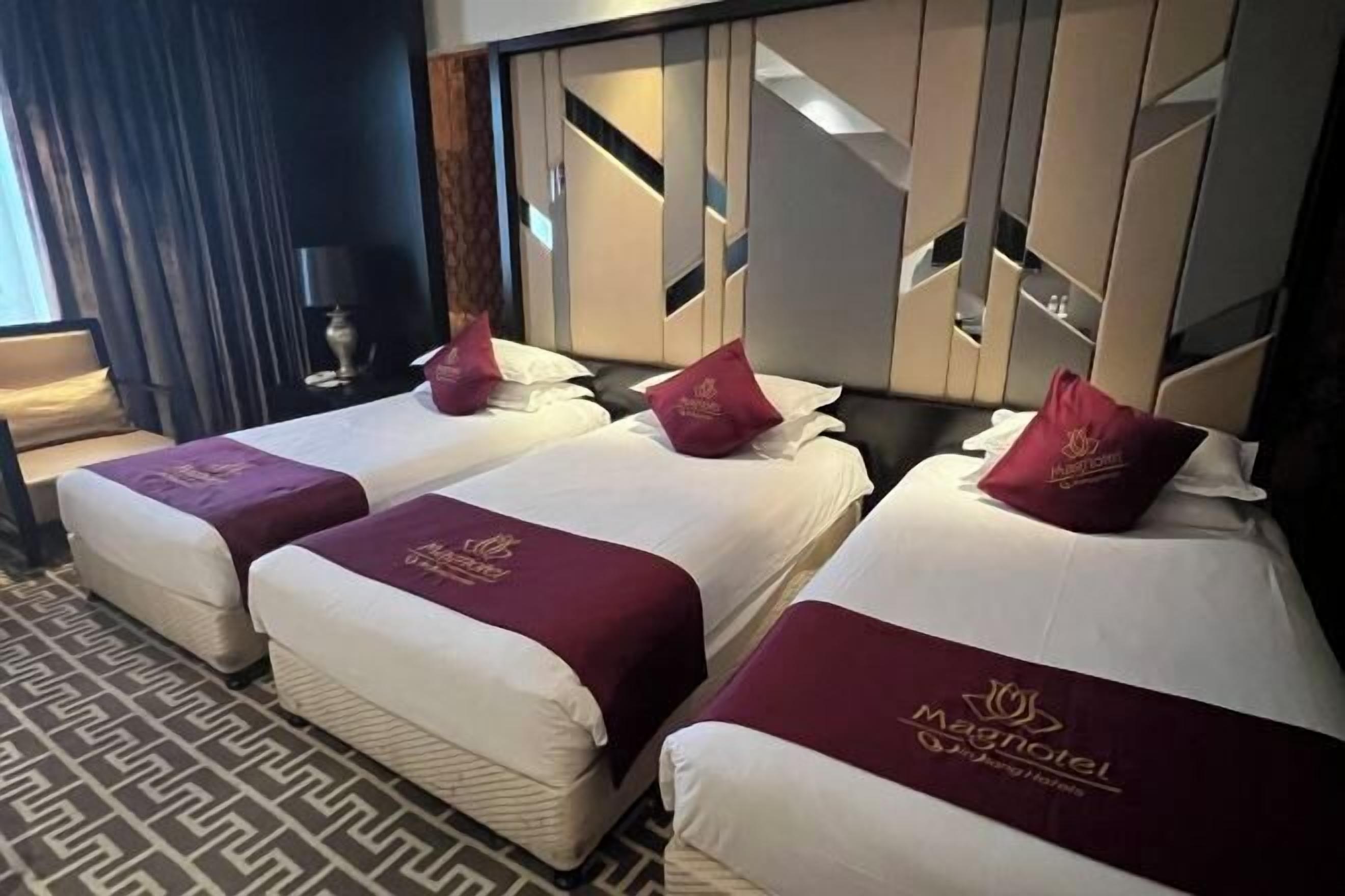 A hotel room with three beds, each with a white mattress and maroon headboard. The room has a patterned carpet and a large mirror on the wall.