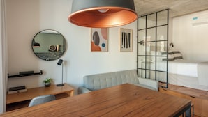 Une chambre d’hôtel moderne avec une table à manger en bois, un miroir rond, un canapé, un lit avec un coussin à rayures et une grande fenêtre.