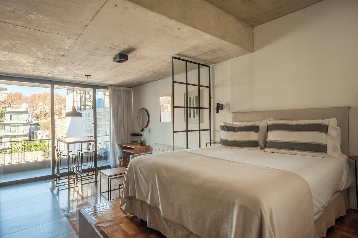 Ein Schlafzimmer mit einem großen Bett, einem Schreibtisch und einem Balkon mit Blick auf Gebäude.