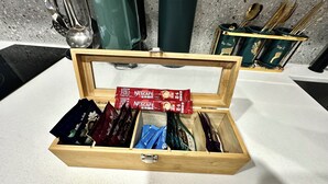 Een houten doos met vakken, gevuld met diverse voorwerpen, waaronder Nescafe koffiehoeveelheden en andere kleine verpakkingen.