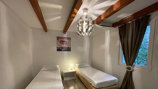 Una habitación con dos camas individuales, una mesita con lámpara, un candelabro en el techo, una ventana con cortinas e imagen colgada en la pared.