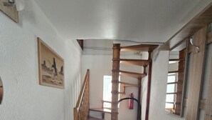 Un estrecho pasillo con una escalera de madera, una silla negra, un escritorio de madera con una revista y un cuadro en la pared.