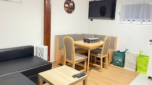 Una sala de estar acogedora con un sofá negro, una mesa de comedor de madera con cuatro sillas, un televisor montado en la pared y una ventana con cortina blanca.