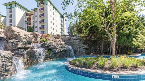 A swimming pool with a waterfall feature, surrounded by a landscaped area with a tree and a stone wall.