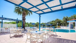 A covered outdoor area with tables and chairs, a pool, and a palm tree. - Muses Hotel (Rhodes)