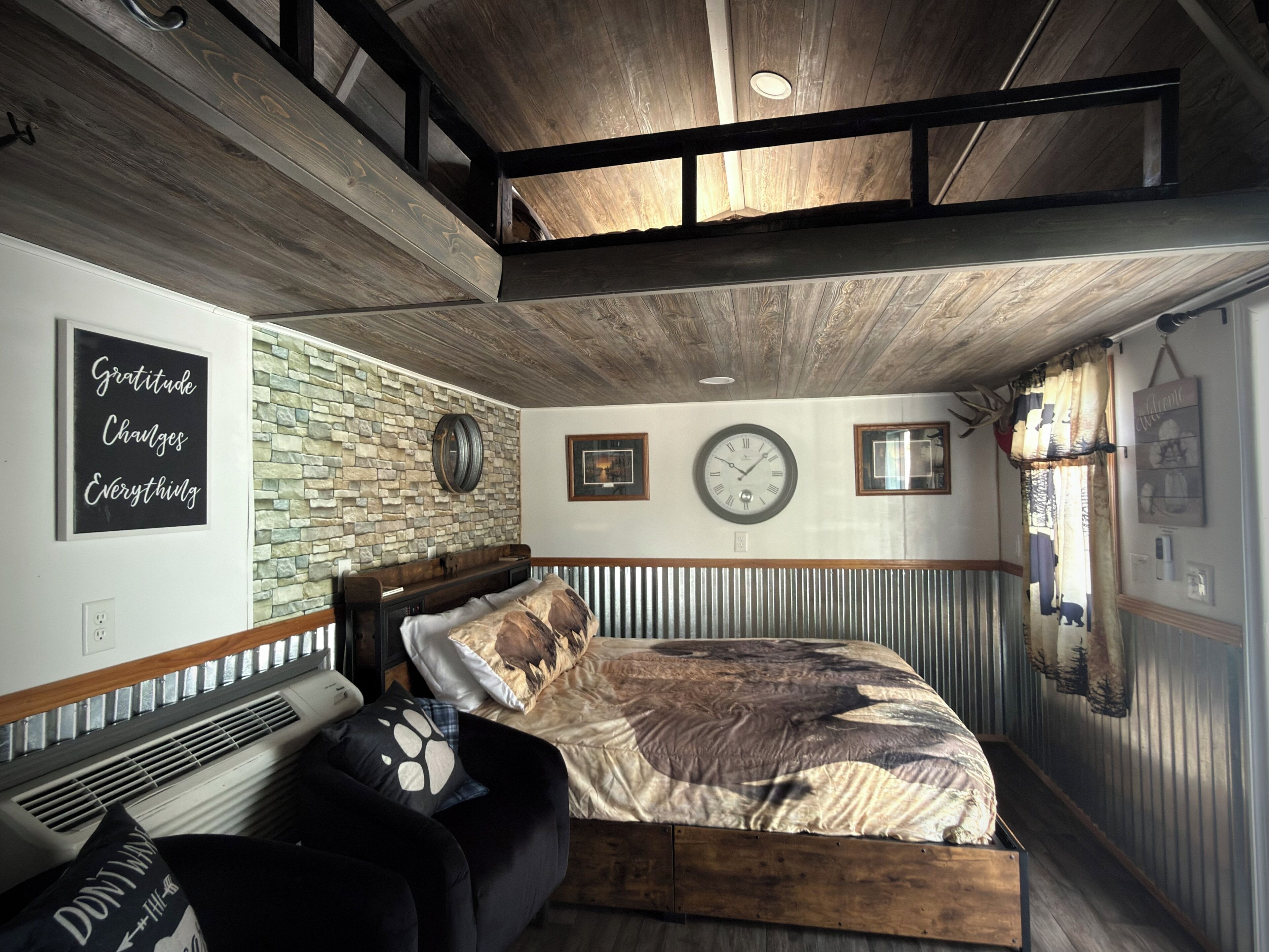 A bedroom with a wooden ceiling, a stone wall, a bed with patterned bedding, a clock, and a motivational quote on the wall.