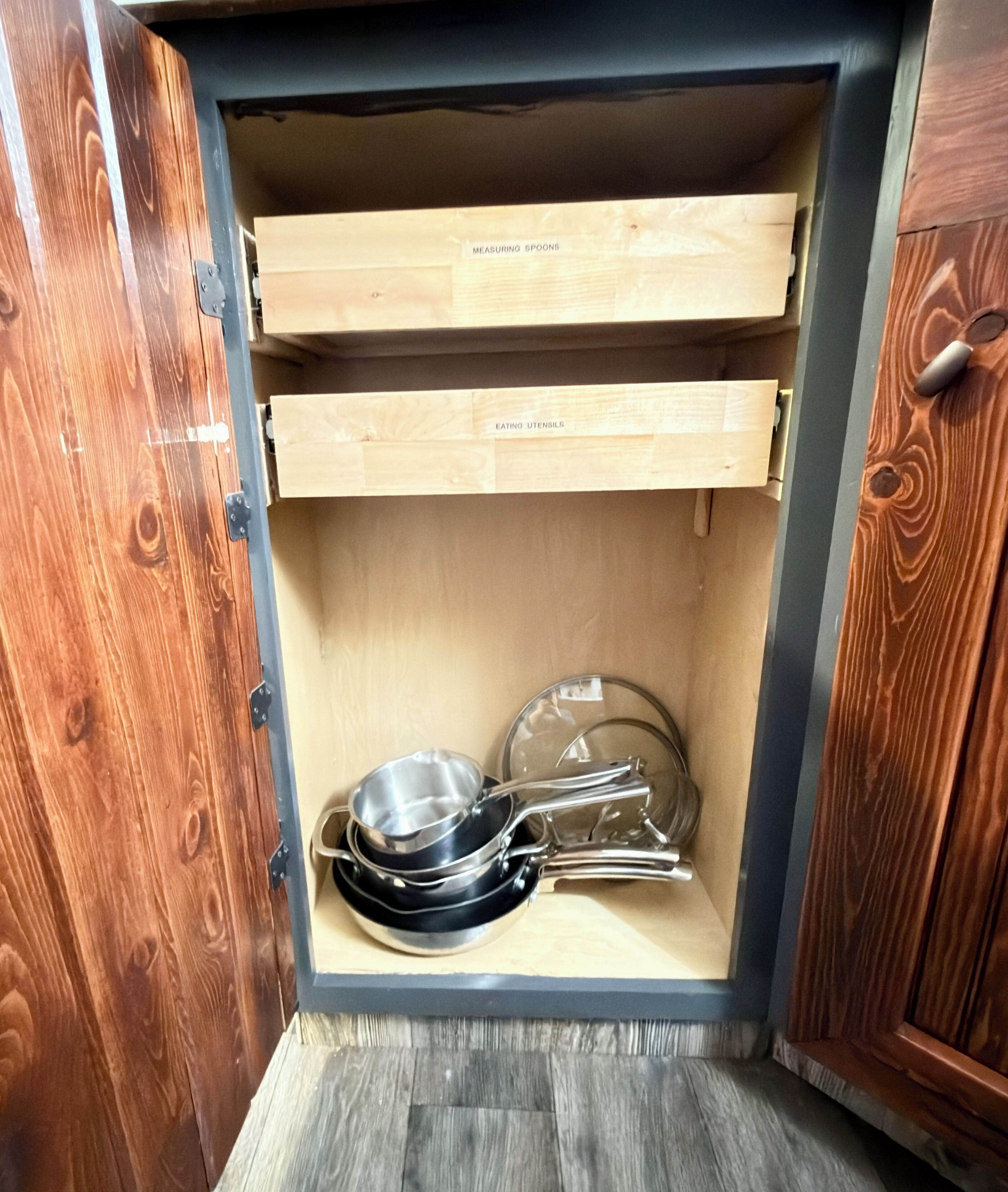 Un mueble de cocina con dos cajones de madera etiquetados 'MEASURING SPOONS' y 'EATING UTENSILS'.