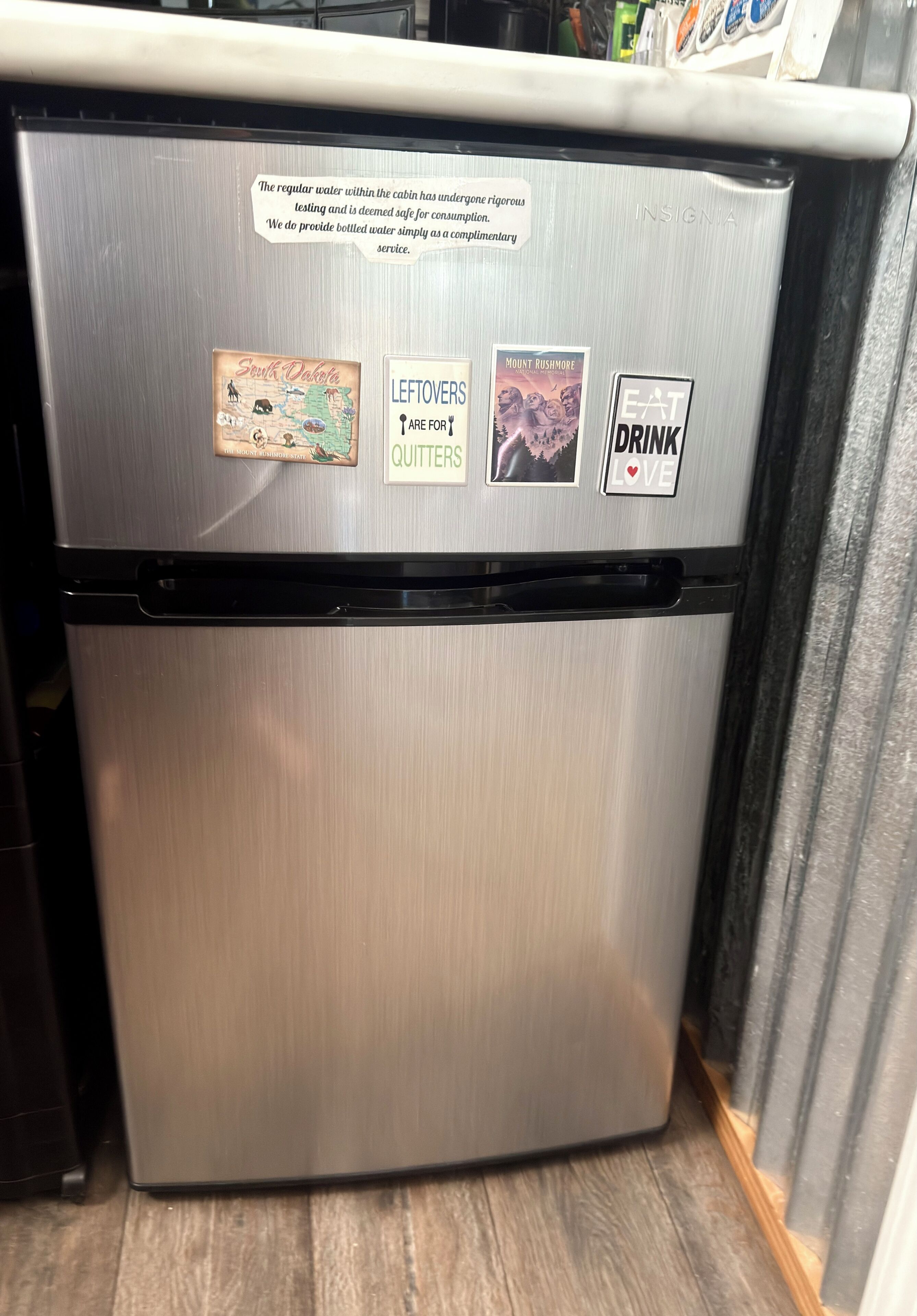 Ein Edelstahl-Kühlschrank mit einem Hinweis zur Wasserqualität, einer Karte von South Dakota, einem Poster mit dem Mount Rushmore und einem Schild mit der Aufschrift: Essen, trinken und lieben.