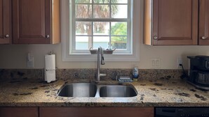 A kitchen with granite countertops, a double sink, wooden cabinets, and a window with a view of palm trees.