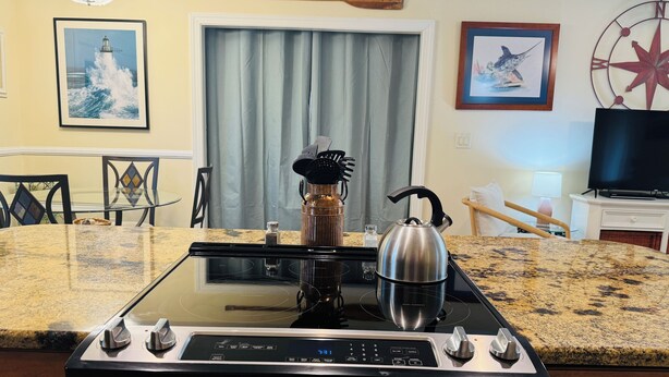 A kitchen with a modern stove, a kettle, and a coffee maker on the counter. There is a dining table with chairs, a television, and artwork on the walls.