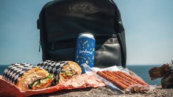 A backpack with a can of Landwash beer, a wrapped sandwich, and a package of sausages on a rocky surface.