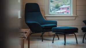 A room with a large window, a blue armchair, a matching ottoman, and a wooden table. - Mallard Inn (St. John's)