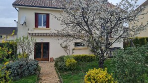 Une maison à deux étages, avec un toit recouvert de tuiles, une antenne parabolique et un jardin agrémenté de diverses plantes ainsi qu’un chemin pavé.