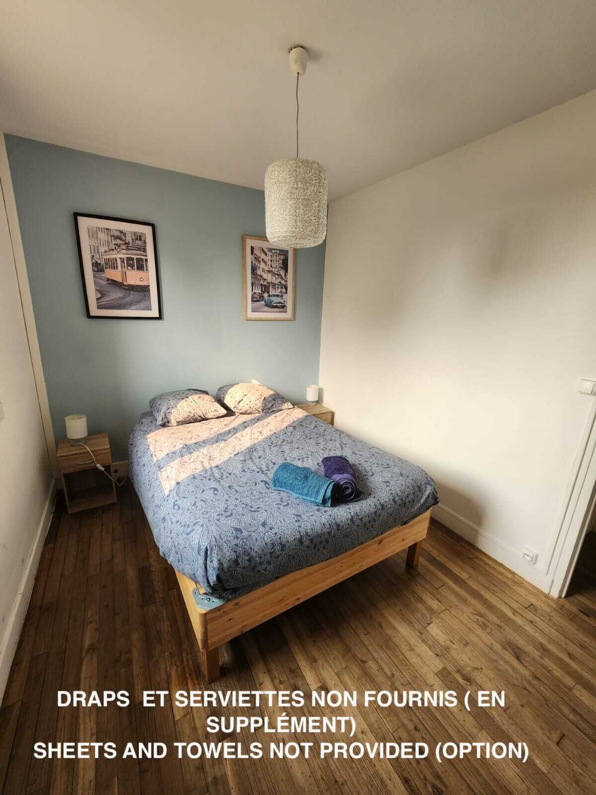 Une chambre avec un lit, deux tableaux accrochés au mur, un luminaire suspendu et un plancher en bois. Le texte indique que les draps et les serviettes ne sont pas fournis.