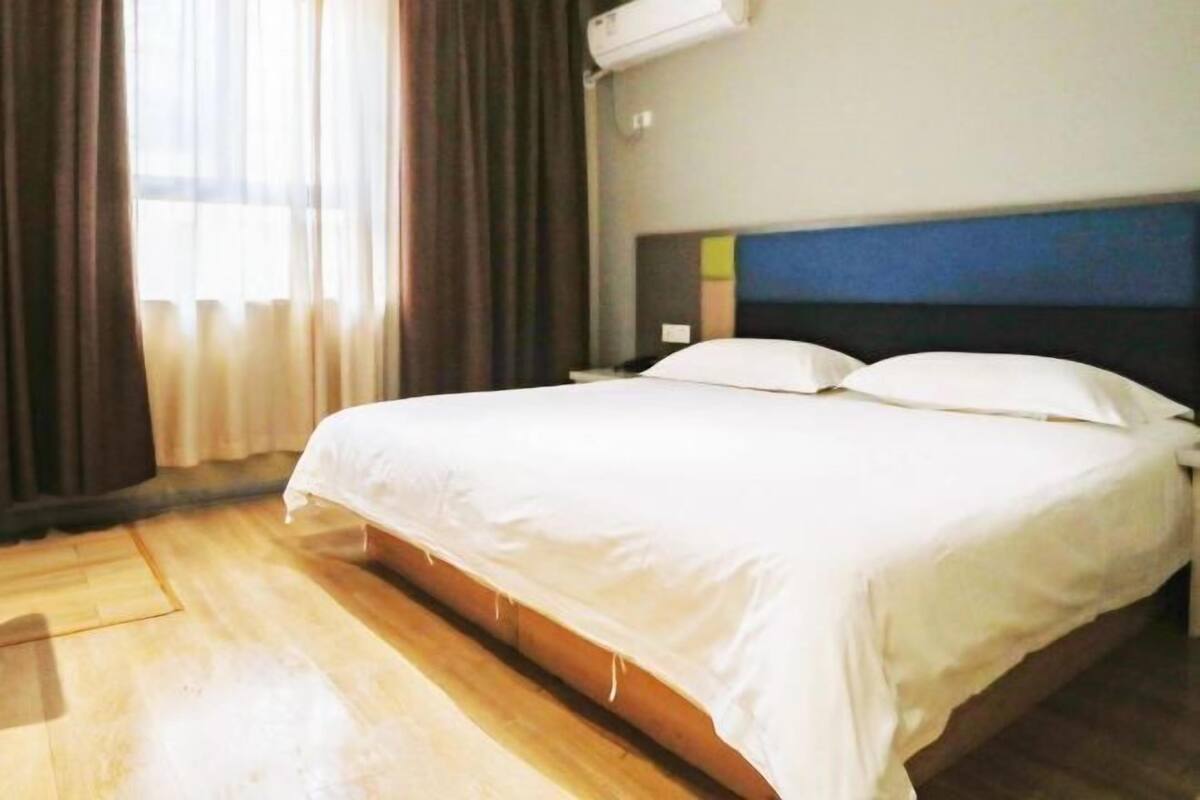 Ein Hotelzimmer mit Bett, Holzboden, einem fenster mit Vorhängen und einer Wand-Klimaanlage.