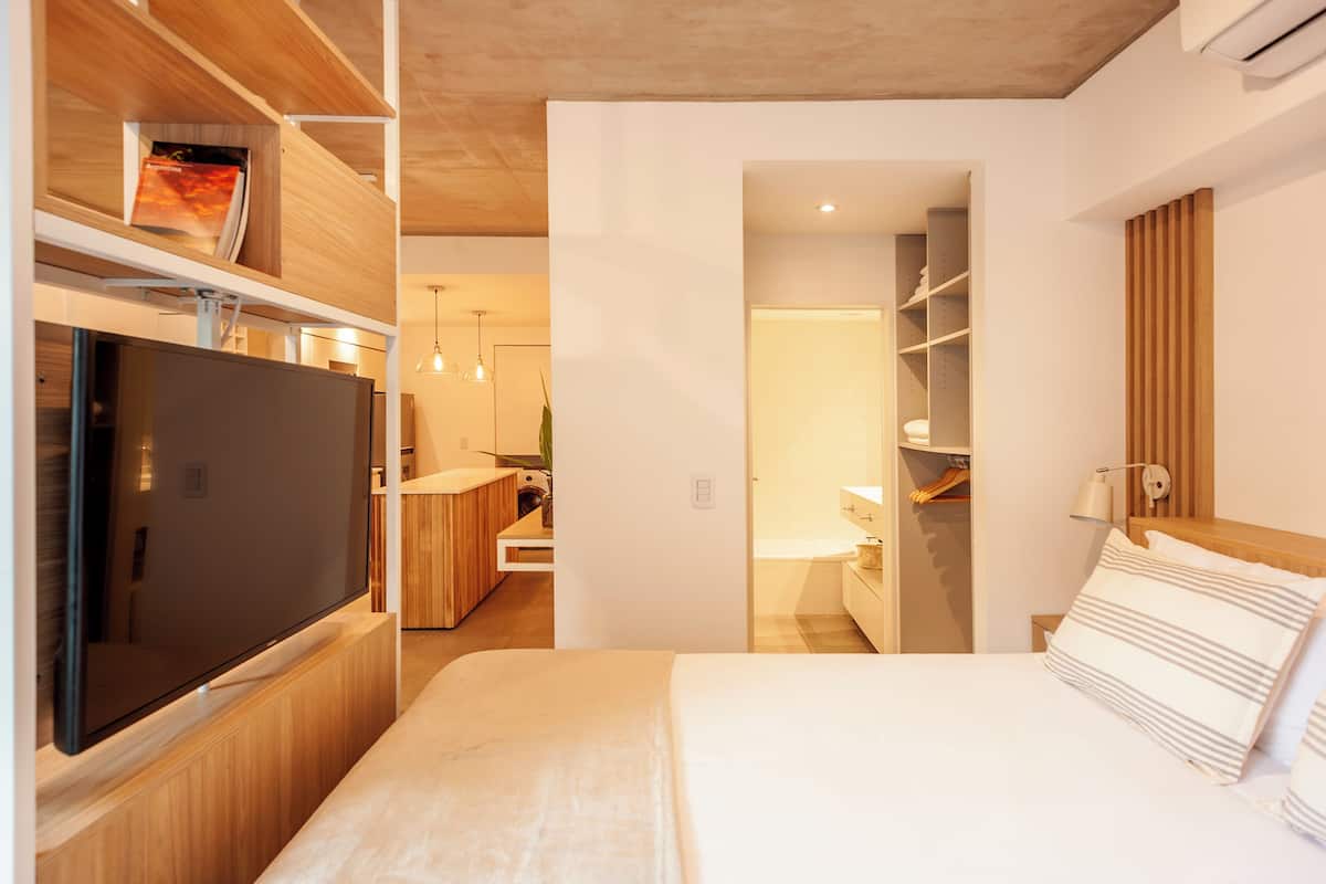 A modern hotel room with a large flat-screen TV, wooden shelving, a bed with striped pillows, and a walk-in closet.