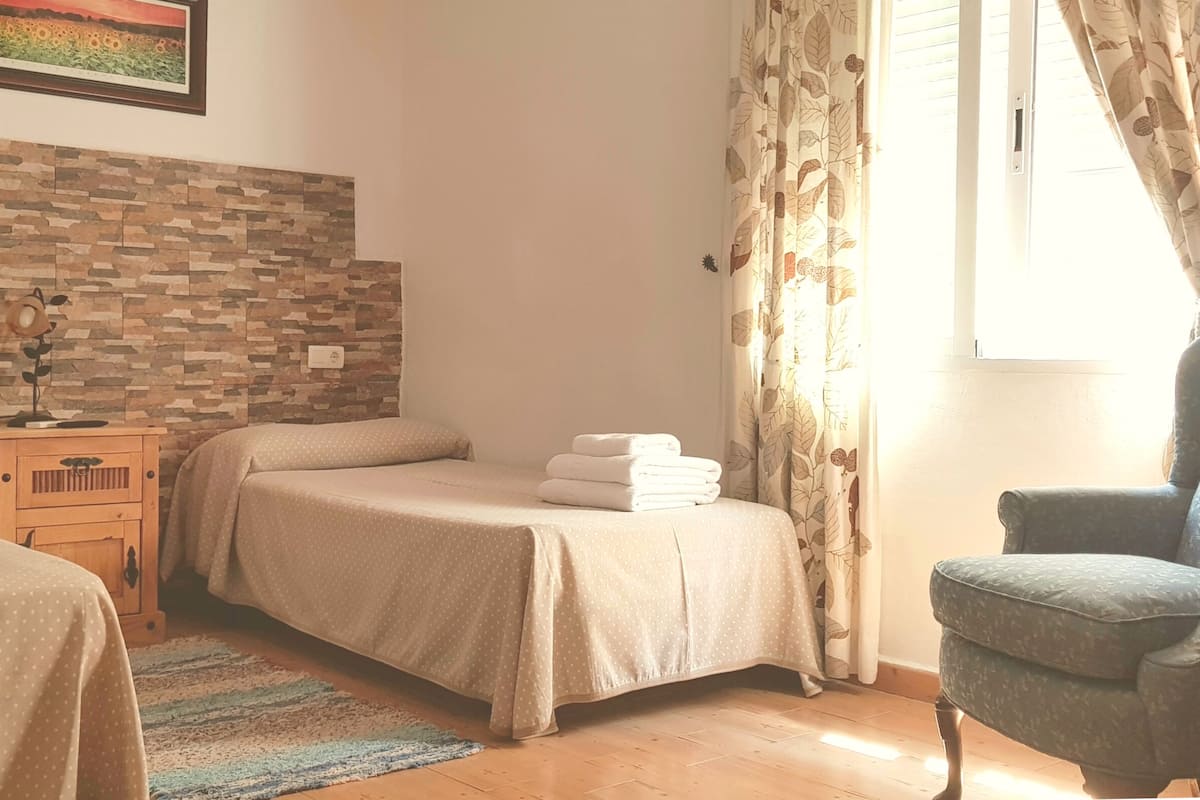 A bedroom with a stone-patterned wall, a bed with a beige bedspread, a wooden nightstand, a chair, and a window with curtains.