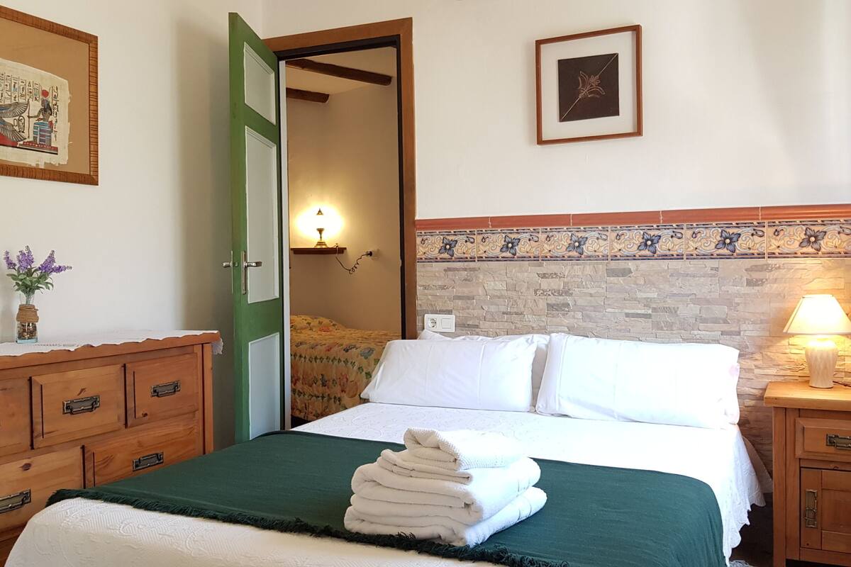 A bedroom with a green bedspread, a stack of white towels, a wooden dresser, a nightstand with a lamp, and a wall-mounted picture.
