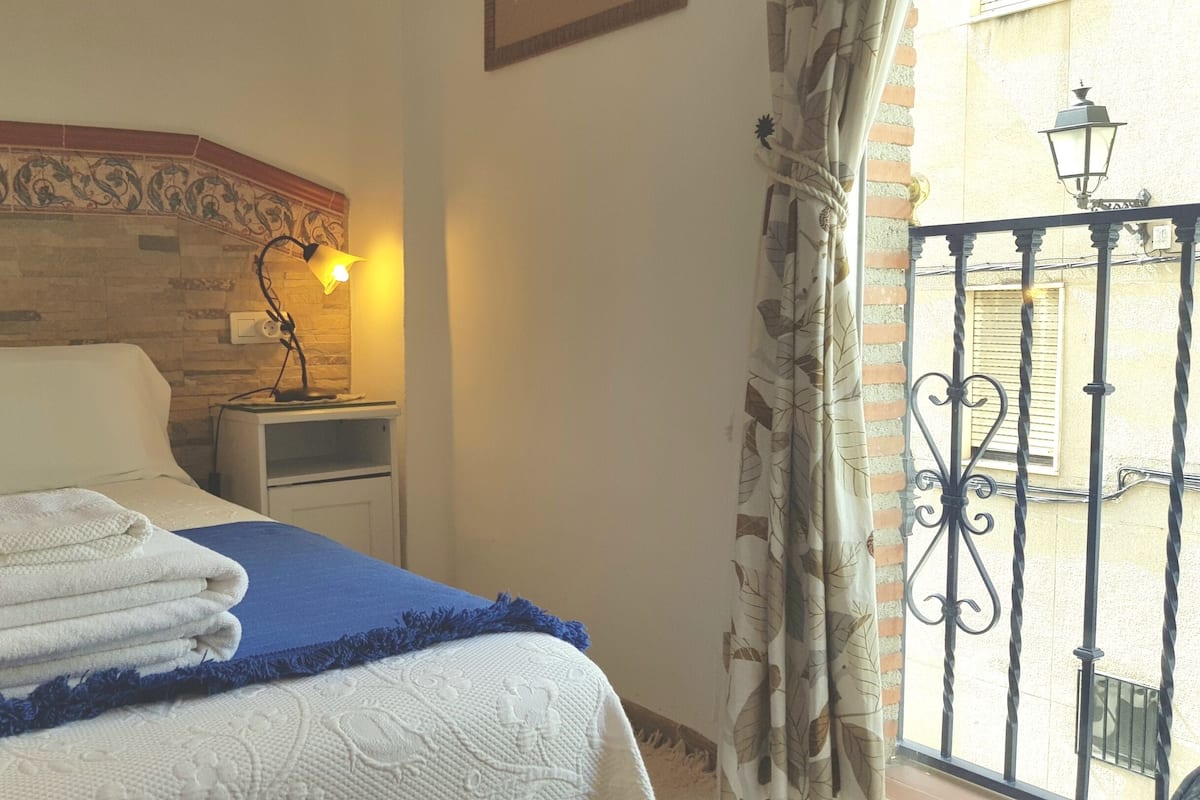 A neatly made bed with folded towels, a bedside lamp, and a view of a balcony with a wrought iron railing.